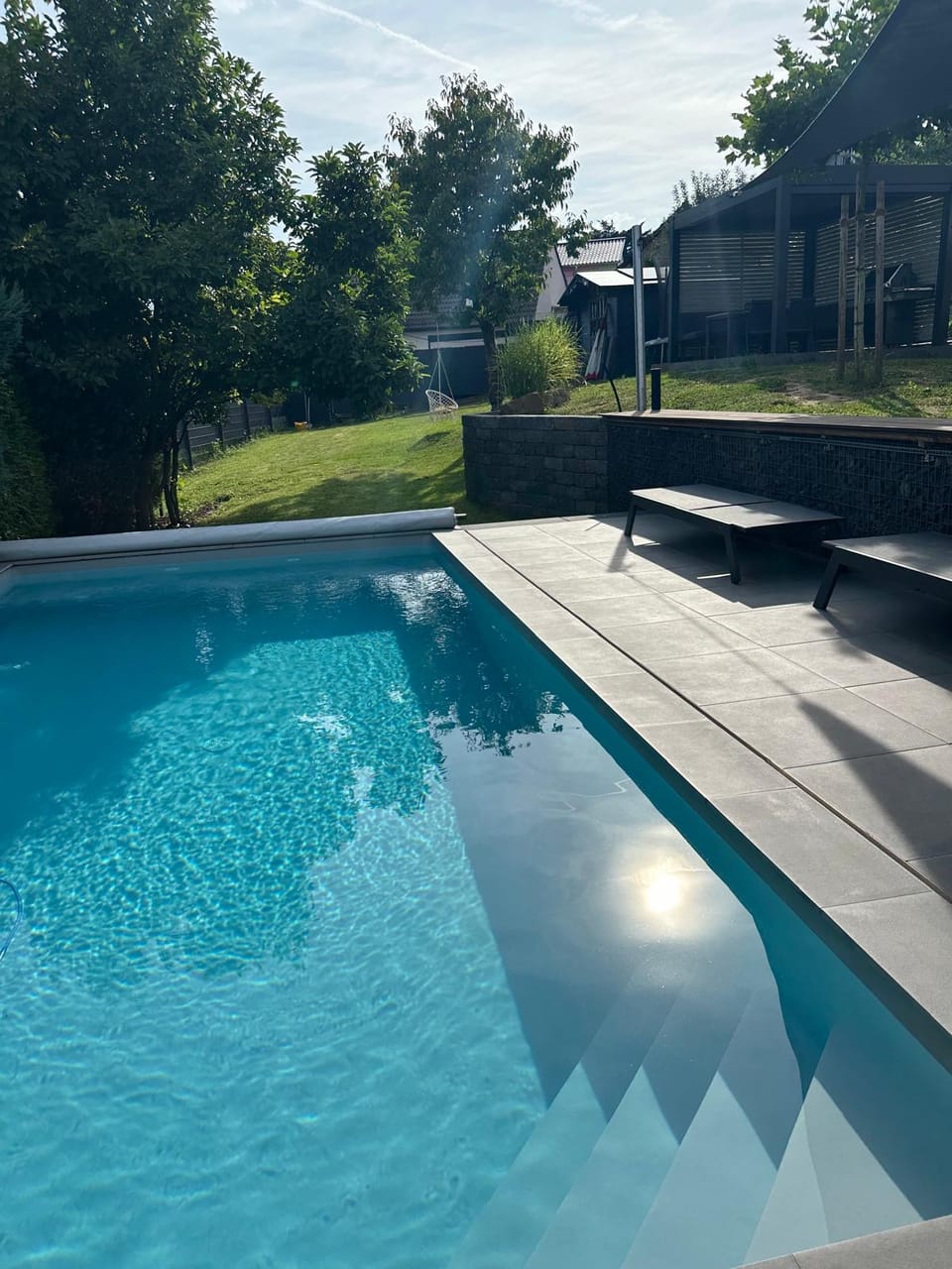 Pool mit Einstiegstreppe, grauen Terrassenplatten und Sonnenliegen im Garten
